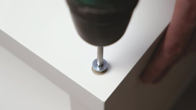 Caucasian man drives hex socket screw drive with electric powered drill or screw gun into white wooden board. Assembling furniture with lag screws at home. Close up