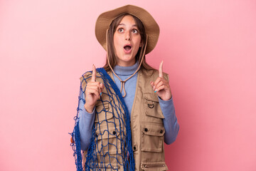 Young caucasian fisherwoman holding a net isolated on pink background pointing upside with opened mouth.