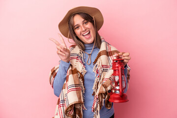 Young caucasian woman holding vintage lantern isolated on pink background joyful and carefree showing a peace symbol with fingers.