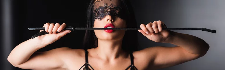 Sensual woman in lace mask and bra holding spanking paddle on black background, banner © LIGHTFIELD STUDIOS