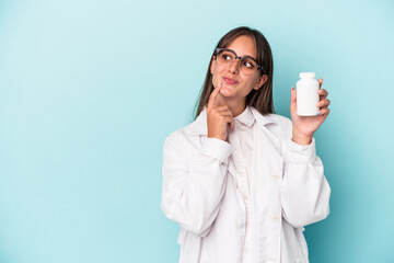 Young pharmacist woman holding pills isolated on blue background looking sideways with doubtful and...