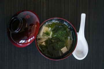 Miso soup in the dark red bowl with lid and white ceramic spoon. Japanese soup in asian restaurant. Vegetarian asian soup with tofu. 