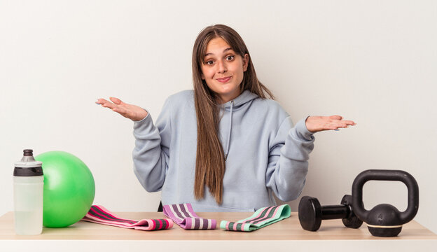 Young caucasian woman sitting at a table with sport equipment isolated on white background doubting and shrugging shoulders in questioning gesture.