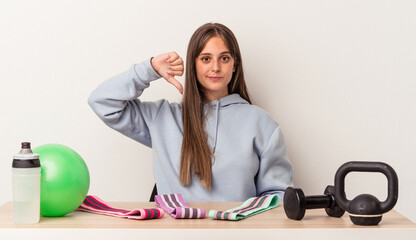 Young caucasian woman sitting at a table with sport equipment isolated on white background showing...