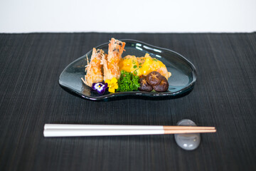 Two shrimps in batter with sauce and mushrooms on the black designed plate. Deep fried prawns decorated with greenery and flower. wooden chopsticks