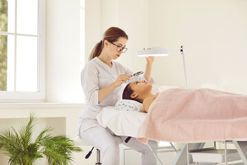 Dermatologist looking at patient's facial skin under loupe and magnifying lamp. Cosmetologist or beautician examining woman's face before removing blackheads, cleaning clogged pores, or curing acne