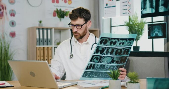 Good-looking concentrated successful professional bearded doctor learning results of x-ray scan and entering datas into computer,filling patient's medical card