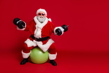 Photo of sportive funny strong old man sit fit ball lift dumbbells wear santa hat costume isolated red color background
