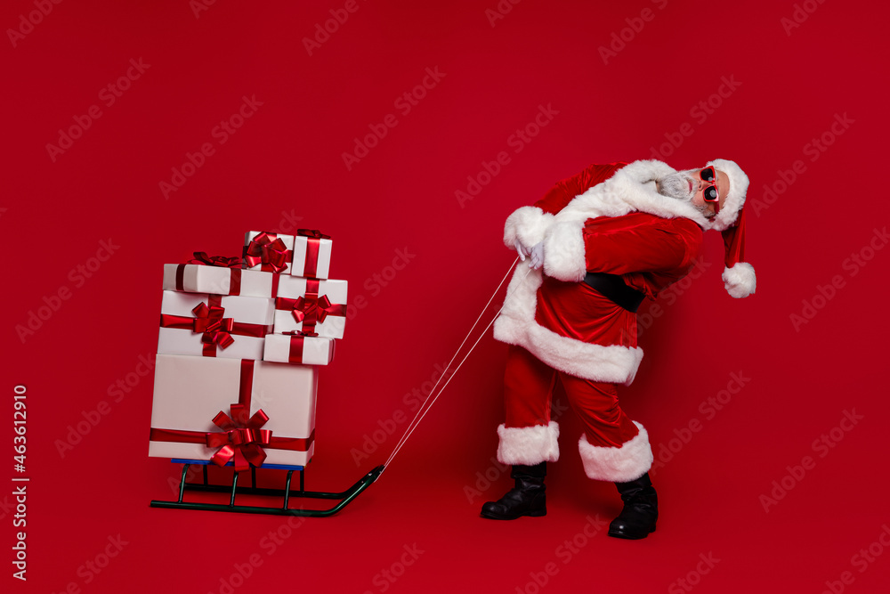 Sticker Photo of strong funky santa claus wear red costume hat dark eyewear dragging sledge presents stack isolated red color background