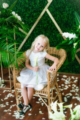  Little cute girl with blond hair in a white dress  and white flowers, lilies and orchids on a  background with a green tropical plants