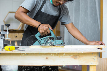 A young male carpenter working using electric planer on wooden plank on his workshop wearing safety equipment