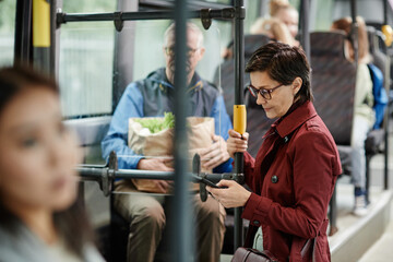 Fototapeta na wymiar Peek shot of elegant adult woman holding onto railing in bus while traveling by public transport in city and using smartphone, copy space