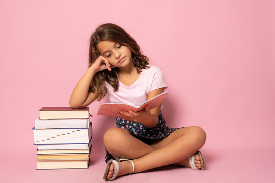 Pretty little girl sit on floor and reading book. Isolated on pink background