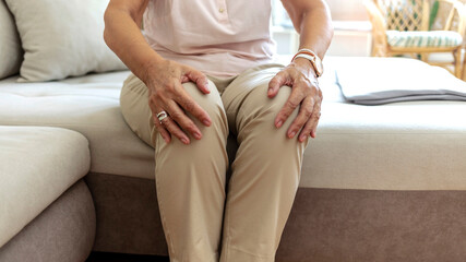 Fototapeta na wymiar Senior woman holding the knee with pain. Elderly woman suffering from pain in knee at home. Old lady touching knee, sitting on sofa, pain in joints, problem with knees. Old age, health problem concept
