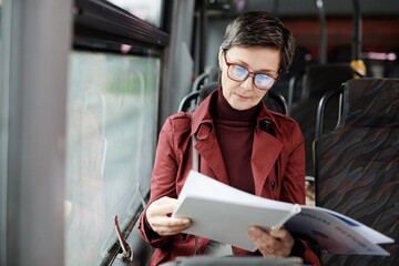 Portrait of elegant mature woman reading book on bus while traveling by public transport in city,...