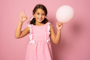 Little smiling girl holding balloon with okay sign isolated over pink background. Celebration concept.