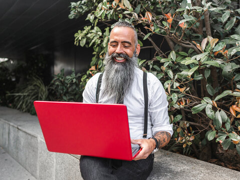 Cheerful bearded businessman typing on laptop on street