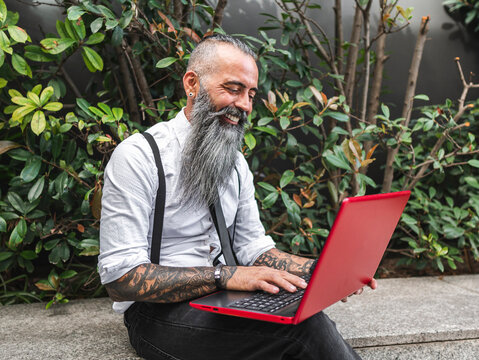 Cheerful bearded businessman typing on laptop on street