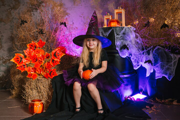 Little cute blonde girl, dressed as a witch on a gloomy background, holds a pumpkin. Decorations for All Saints Day. Halloween costume.