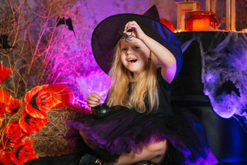 Little cute blonde girl, dressed as a witch on a gloomy background, holds a spider in her hand....
