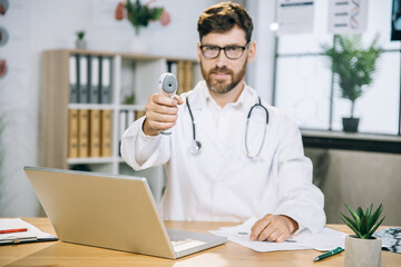 Blur view of Caucasian medical specialist posing on camera while sitting at table with modern contactless thermometer in hands. Selective focus on thermometer in hands
