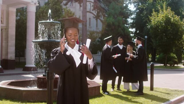 In a graduation day in the college garden a pretty young black woman have a discussion on the phone after she was graduate other students multiracial discussing in group on the background
