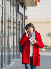 Woman in red duffle coat talks by smartphone. Woman walks down the street grasping cup of take away coffee and paper cheque.