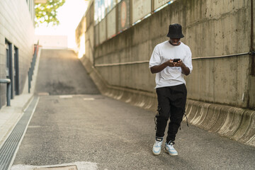 Chico negro atlético posando con el smartphone en una calle gris con ropa urbana