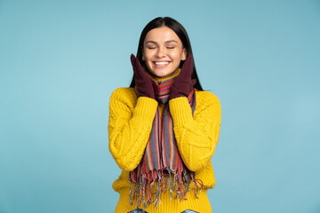 Smiling gorgeous woman in stylish yellow sweater closed her eyes while standing in front of the camera and posing on blue wall background. Concept of winter holidays