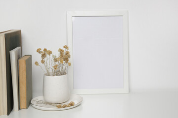 Horizontal white frame mockup on table with books, ceramic vase with yellow dry Everlasting flowers. White wall background.