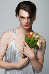 young transgender man in slip dress holding flowers isolated on gray
