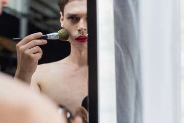 young transgender man applying face powder with cosmetic brush near mirror
