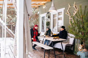 Friends sitting on a snowy house terrace under a roof, talking to each other, drinking coffee....