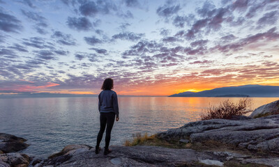 Caucasian Woman on a rocky coast during a dramatic cloudy sunset. Lighthouse Park ,Horseshoe Bay, West Vancouver, British Columbia, Canada. Adventure Travel Concept