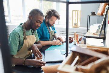 Portrait of two modern male artisans working while sitting by desk in leatherworkers workshop, copy...