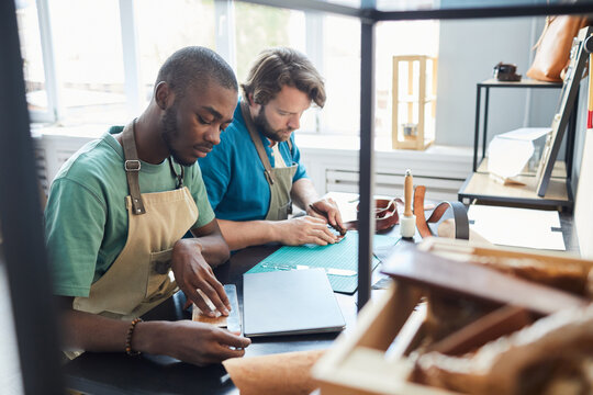 Portrait of two modern male artisans working while sitting by desk in leatherworkers workshop, copy space