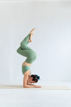Young woman in green sports bra and leggings practices yoga at home. Balance exercise. Spiritual development. Variation of vrischikasana pose.