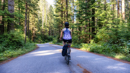 Fototapeta premium Adventurous White Caucasian Woman on a bicycle riding on a path in Green Canadian Rain Forest. Seymour Valley Trailway in North Vancouver, British Columbia, Canada.