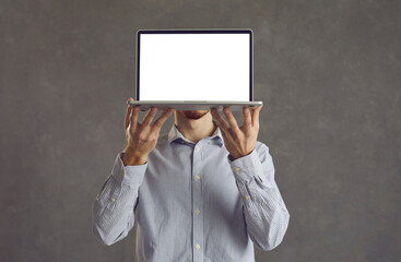 Unrecognizable man holding his laptop with white blank screen at face height while standing on gray background. Young businessman hides his face behind computer showing copy space. Banner Advertising.