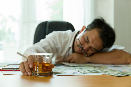 Stressed adult Asian businessman at workplace and drinking alcohol with many document paper for business on desk. Asian man overstressed at job while sitting at office desk in the office