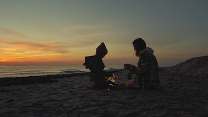 Mid shot of a mother and her daughter celebrating on the beach with a glowing garland in their arms and hot tea in thermos at sunset. Holiday atmosphere at the seaside.
