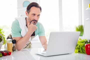 Photo of focused concentrated man look screen netbook cook lesson wear apron blue t-shirt home kitchen indoors