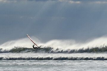 Windsurfer sur une vague pleine d'embruns sous les rayons du soleil qui percent à travers les nuages