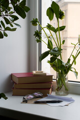 Interior home scene. A stack of old books, writing materials and a bouquet of flowers on the windowsill against the background of the window.