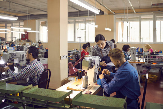 People working in a big workshop room at a shoe factory. Male and female workers sitting at tables with industrial sewing machines and making new footwear details. Manufacturing industry concept