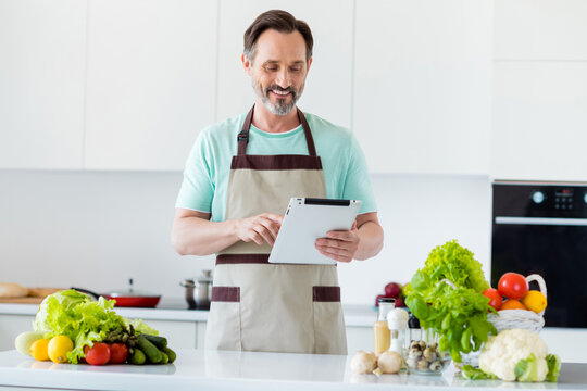 Photo of modern mature man hold tablet choose recipe typing wear apron blue t-shirt home kitchen indoors