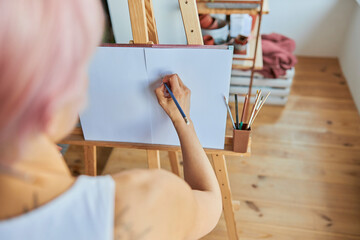 Woman painter uses graphite pencil to draw picture on blank paper at wooden easel