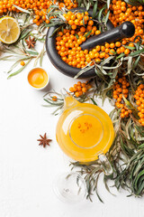Sea buckthorn hot tea in a glass teapot with lemon and spices, branch of fresh, juicy, ripe yellow berries on white background. Vitaminic healthy tea. Hot winter drink. Top view
