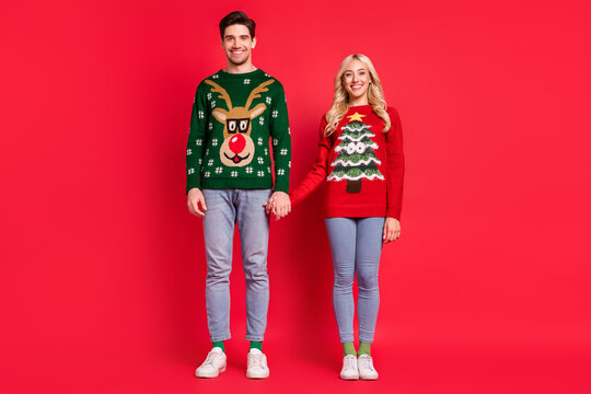 Photo of cheerful positive couple hold hands toothy smile wear ugly sweater jeans shoes isolated red color background