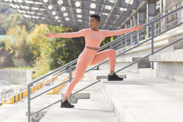 Fototapeta na wymiar Female athlete doing fitness in a pink sports suit near the sports stadium, African American performs fitness exercises and stretching in the morning